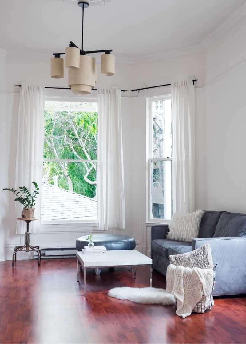 Salón luminoso con dos ventanales de guillotina, cortinas blancas, sofá gris y suelo de madera rojiza