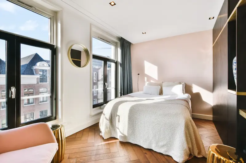 Dormitorio de un piso turístico con cama vestida en blanco, suelo de madera en espiga y ventanales a la calle