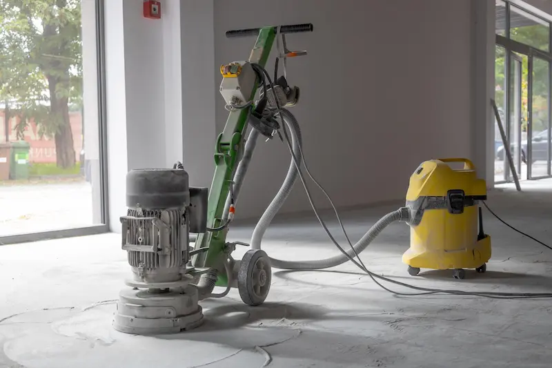 Pulidora de suelos y aspirador industrial amarillo sobre un suelo con polvo de obra junto a un ventanal