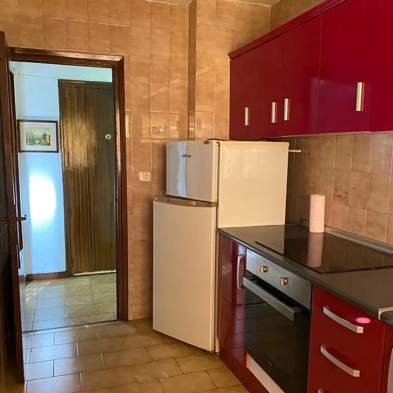 Cocina estrecha con muebles rojos brillantes, frigorífico blanco, placa negra y suelo de baldosa cerámica